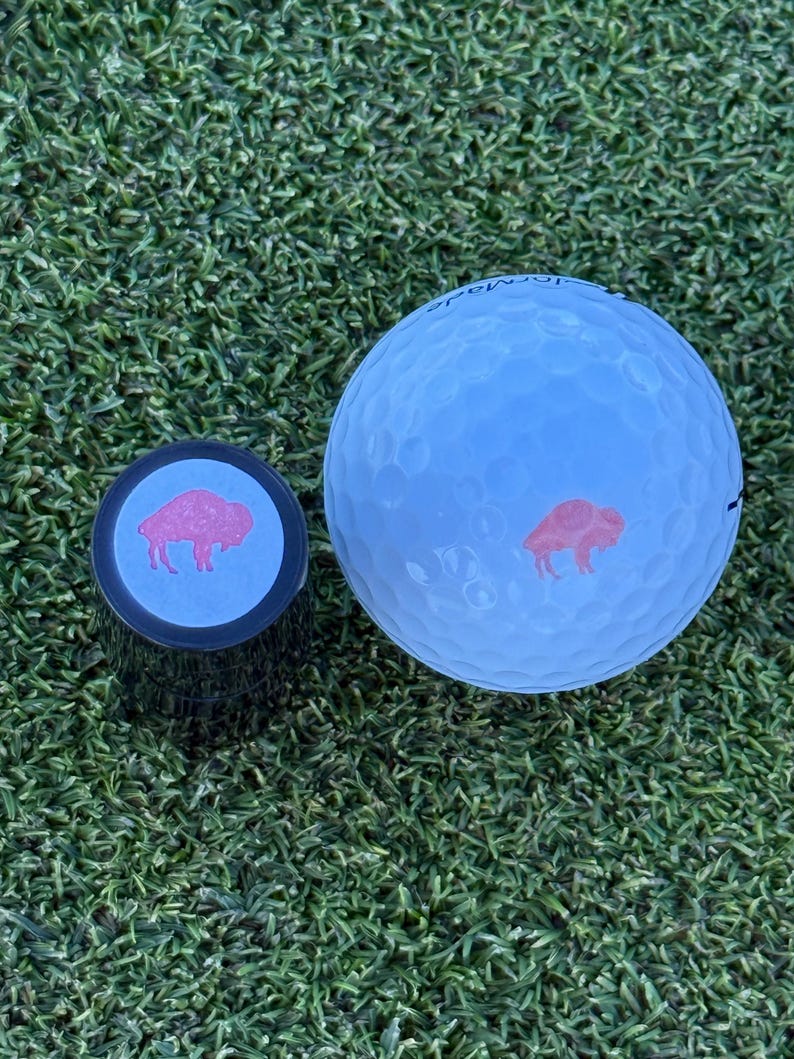 May include: A white golf ball with a red buffalo logo and a black golf ball marker with a blue background and a red buffalo logo.