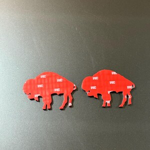 May include: Two red buffalo-shaped stickers with 3M adhesive backing. The stickers are cut in the shape of a buffalo, with the adhesive visible on the back. The stickers are on a gray surface.