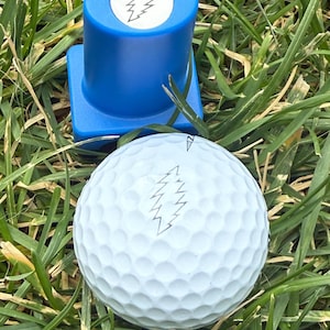 May include: A white golf ball and a blue golf ball marker with a lightning bolt design are set on a bed of green grass. The golf ball has the same lightning bolt design. The golf ball is dimpled.
