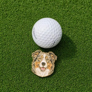 May include: A white Titleist golf ball with a dimpled surface positioned above a metal enamel pin featuring a detailed illustration of a brown, white, and grey Australian Shepherd dog face with its tongue out and mouth slightly open.