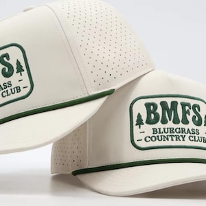 May include: Two off-white baseball caps with perforated side panels and a dark green rope detail across the brim. Each cap features a rectangular patch with dark green text reading BMFS BLUEGRASS COUNTRY CLUB and two small pine tree icons.