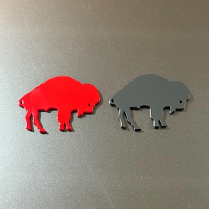 May include: Two bison-shaped cutouts. One is bright red, the other is dark gray. Both have a smooth, flat surface and are placed on a gray background. The bison are facing left, with their legs and heads clearly defined.