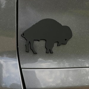 May include: A black bison emblem affixed to a gray vehicle. The emblem is a silhouette of a bison, a large, shaggy-haired animal, and is mounted on the rear of the car. The bison is facing right.