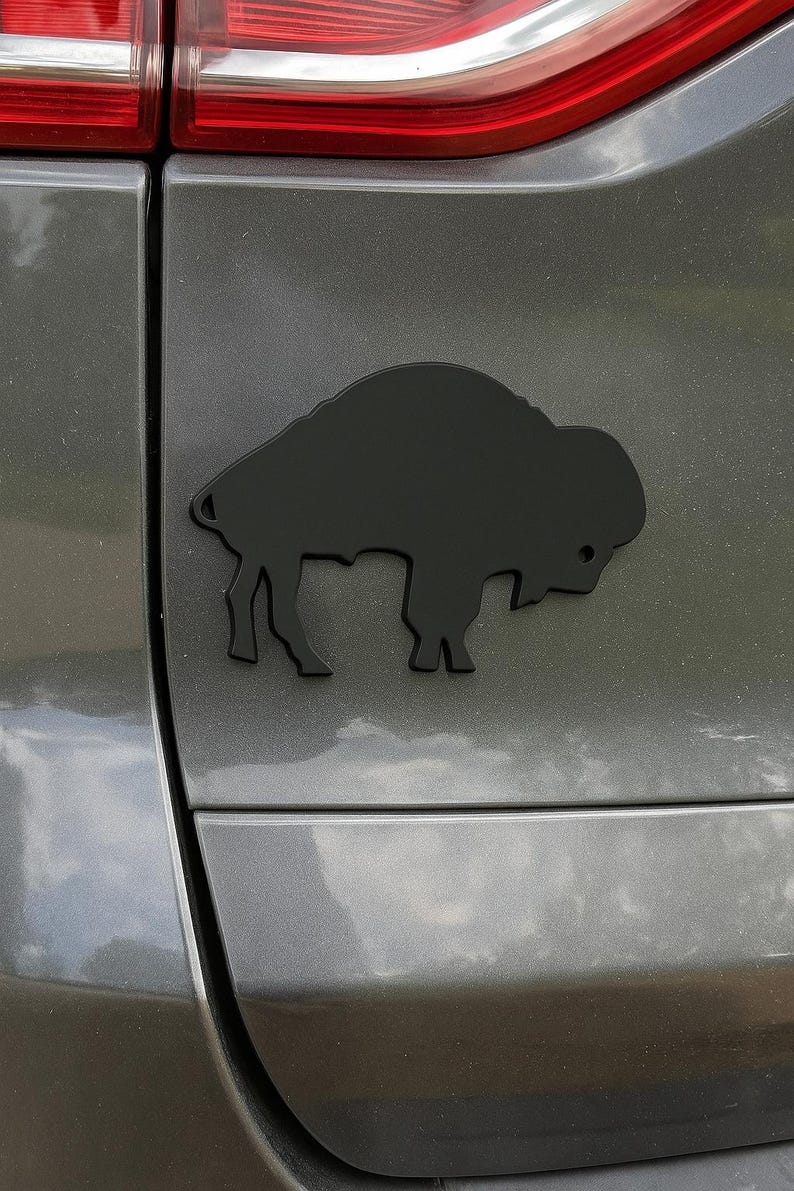 May include: A black bison emblem affixed to a gray vehicle. The emblem is a silhouette of a bison, a large, shaggy-haired animal, and is mounted on the rear of the car. The bison is facing right.
