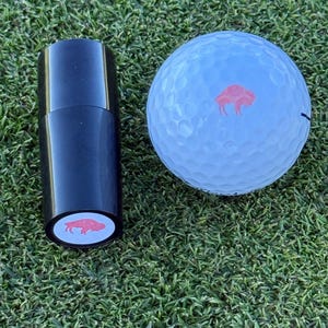 May include: A black golf ball marker with a red buffalo silhouette on a white background. The marker is next to a white golf ball with a red buffalo silhouette on a white background.