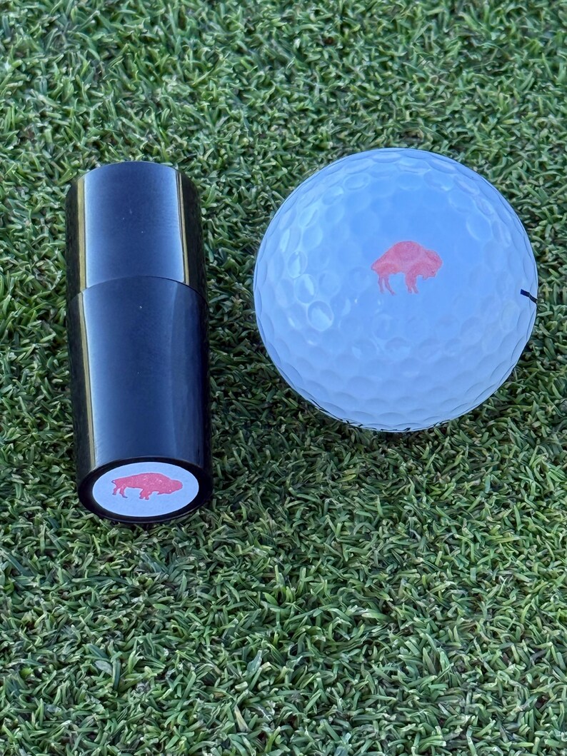May include: A black golf ball marker with a red buffalo silhouette on a white background. The marker is next to a white golf ball with a red buffalo silhouette on a white background.
