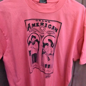 May include: A pink t-shirt with a black graphic print of two people holding guns. The graphic says "Grand American Trapshooting Legends".