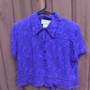 May include: A purple button-up blouse with a floral pattern and a lace trim. The blouse has a collar and short sleeves. The label reads "Karen Stevens Petite."