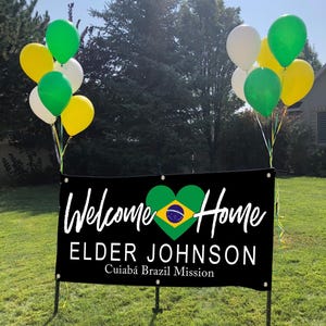 a banner with the text "Welcome Home Elder Johnson" and "Cuiabá Brazil Mission" displayed on it, along with a heart symbol. The banner is set against a backdrop of trees and balloons, creating a festive atmosphere.
