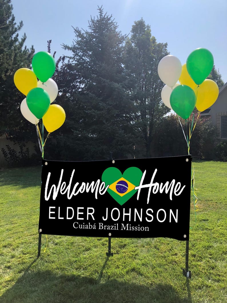 a banner with the text "Welcome Home Elder Johnson" and "Cuiabá Brazil Mission" displayed on it, along with a heart symbol. The banner is set against a backdrop of trees and balloons, creating a festive atmosphere.