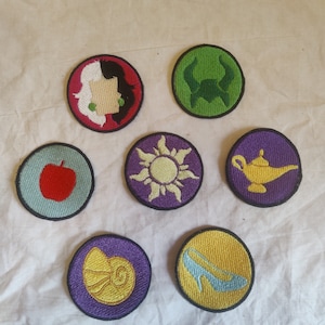 Può includere: Sette toppe ricamate da stirare con personaggi Disney. Le toppe sono circolari e presentano vari colori, tra cui viola, verde, rosso, giallo e blu. I personaggi includono un genio, una lampada magica, una mela avvelenata, una scarpetta di cristallo da principessa, una conchiglia, un sole e un cattivo con i capelli neri.