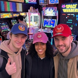May include: Three baseball caps featuring character graphics: a blue cap with a circular Sonic the Hedgehog patch, a pink cap with an Amy Rose patch, and a red cap with a Knuckles the Echidna patch. Each cap has a structured crown and a curved brim.