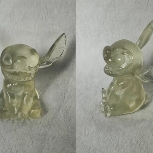 Puede incluir: Una figurita de Stitch azul transl&uacute;cida con orejas grandes y una amplia sonrisa. La figurita est&aacute; sentada con las piernas cruzadas y los brazos cruzados.