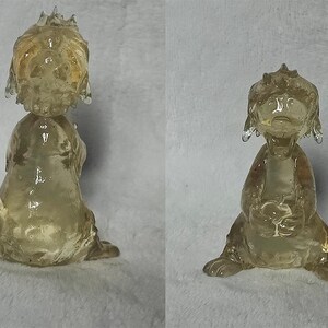 Puede incluir: Una figurita de vidrio transparente de un perro de dibujos animados con un tinte marr&oacute;n. El perro est&aacute; sentado sobre sus patas traseras con las patas delanteras levantadas. La figurita mide aproximadamente 15 cm de alto.