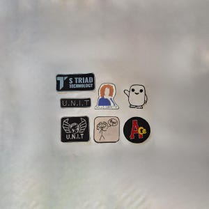 Può includere: Una collezione di sette toppe da stirare. Le toppe sono di varie forme e dimensioni e presentano diversi disegni, tra cui un personaggio dei cartoni animati, un fantasma, un logo con il testo "S Triad Technology", un logo con il testo "U.N.I.T" e un logo con il testo "Ace".