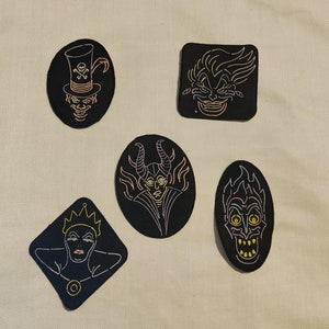 May include: Five black and white embroidered iron-on patches featuring Disney villains. The patches feature Maleficent, Ursula, Cruella de Vil, the Evil Queen, and Hades.