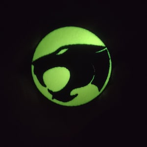 May include: A round, neon green patch with a black silhouette of a panther's head. The panther is inside a circle, and the background is black. The patch has a fuzzy texture.