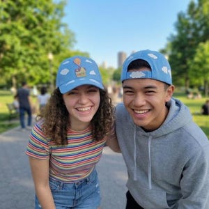 May include: Two light blue baseball caps featuring a white cloud pattern across the fabric. One cap displays a small embroidered patch of a house attached to a cluster of colourful balloons on the front panel, while the other cap has a cloud design.