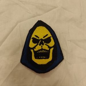 May include: A yellow and black embroidered patch of a skull wearing a blue hooded robe.