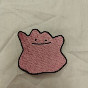以下が含まれることがあります： ピンクと黒の刺繍パッチ。笑顔のピンクのブロブの漫画。