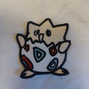 May include: White and black embroidered patch of a Pokemon character, a white Eevee with red and blue spots, emerging from an egg.