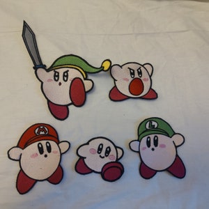 May include: Five embroidered patches featuring characters from the video game Kirby. The patches are pink, red, green, and white with black outlines. The characters are wearing different outfits and accessories, including a sword, a hat, and a crown.