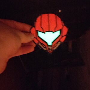 May include: A red and black patch in the shape of a helmet with a glowing green visor. The patch is held against a dark background. The helmet design is reminiscent of a science fiction character.