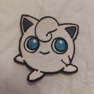 May include: Embroidered patch featuring a cartoon character with a round, light pink body, large blue eyes, and a small smile. The character has dark blue accents on its ears and a curled tuft of hair. The patch is outlined in black.