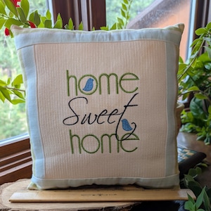 May include: Square decorative pillow with the embroidered phrase "home sweet home" in green and black script. The pillow has a light beige center with a light blue border and two small blue bird designs. A wooden ruler rests in front of the pillow.