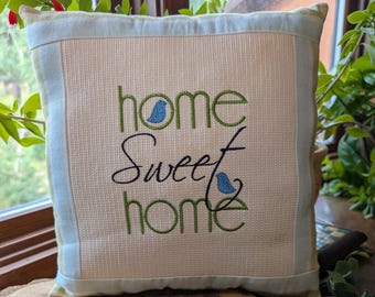 May include: Square decorative pillow with the embroidered phrase "home sweet home" in green and black script. The pillow has a light beige center with a light blue border and two small blue bird designs. A wooden ruler rests in front of the pillow.