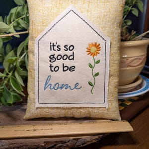 May include: A rectangular yellow textured pillow featuring a house-shaped fabric patch with the embroidered text "it's so good to be home" and a single orange sunflower. A wooden ruler is positioned at the base for scale reference.