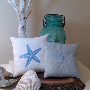 Peut inclure: Deux coussins carr&eacute;s d&eacute;coratifs avec des motifs d'&eacute;toiles de mer brod&eacute;es en bleu et bleu clair. Un grand bocal turquoise transparent avec une poign&eacute;e en m&eacute;tal noir est derri&egrave;re les coussins. Un coquillage et du bois flott&eacute; compl&egrave;tent le th&egrave;me c&ocirc;tier.