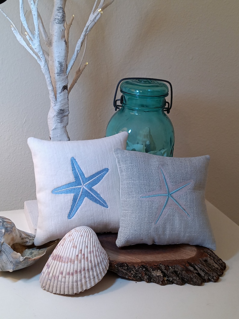 Peut inclure: Deux coussins carr&eacute;s d&eacute;coratifs avec des motifs d'&eacute;toiles de mer brod&eacute;es en bleu et bleu clair. Un grand bocal turquoise transparent avec une poign&eacute;e en m&eacute;tal noir est derri&egrave;re les coussins. Un coquillage et du bois flott&eacute; compl&egrave;tent le th&egrave;me c&ocirc;tier.