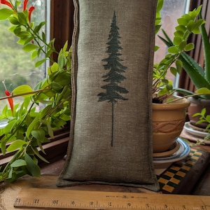 May include: A rectangular sage green linen pillow featuring a dark green embroidered pine tree design centered on the front. A wooden ruler is placed horizontally in front of the pillow, showing a length of approximately 12 inches.