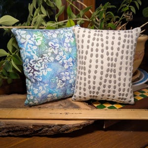 Peut inclure: Deux petits coussins d&eacute;coratifs. L'un pr&eacute;sente un motif floral bleu et blanc. L'autre a un fond blanc avec un motif r&eacute;p&eacute;titif de points gris. Les coussins sont rectangulaires et semblent faits &agrave; la main.