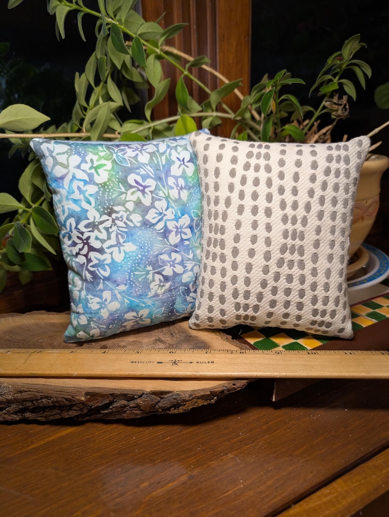 Peut inclure: Deux petits coussins d&eacute;coratifs. L'un pr&eacute;sente un motif floral bleu et blanc. L'autre a un fond blanc avec un motif r&eacute;p&eacute;titif de points gris. Les coussins sont rectangulaires et semblent faits &agrave; la main.