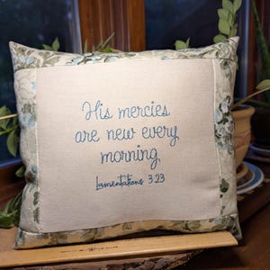 May include: A square throw pillow with a central cream-colored fabric panel featuring blue embroidered text that reads His mercies are new every morning Lamentations 3:23, framed by a border of floral patterned fabric in shades of blue and green.