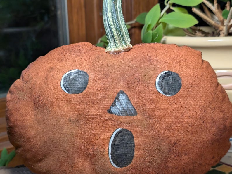 May include: A plush pumpkin decoration made of textured, rust-coloured fabric with a painted face featuring two round black eyes, a triangular nose, and an oval mouth. A realistic, light green, ribbed pumpkin stem is attached to the top.