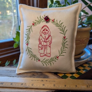 May include: A square off-white fabric cushion featuring red embroidered Santa Claus holding a candle, surrounded by a green embroidered wreath with red berries and a small red jingle bell at the top. A wooden ruler shows the cushion is 12.7 cm wide.