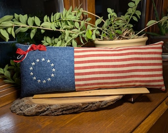 May include: A rectangular throw pillow featuring a blue field with a circle of thirteen white embroidered stars on the left and horizontal red and white stripes on the right. A small red ribbon is tied to the top left corner of the pillow.