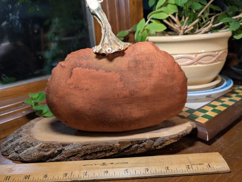 May include: A terracotta-coloured fabric pumpkin with a textured surface and a metallic silver stem, resting on a natural wood slice. A wooden ruler is positioned in the foreground, showing the pumpkin is approximately 15 cm wide.