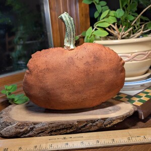 May include: A textured, rust-coloured fabric pumpkin with a light green stem, resting on a natural wood slice. A wooden ruler is placed in front, showing the pumpkin is approximately 15 cm wide at its base on the wood slice.