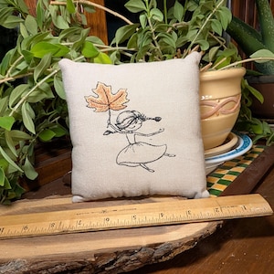 May include: A square, off-white fabric pillow featuring a black embroidered line drawing of a figure holding a large, orange-brown maple leaf. A wooden ruler is placed in front, showing the pillow is approximately 5 inches wide.