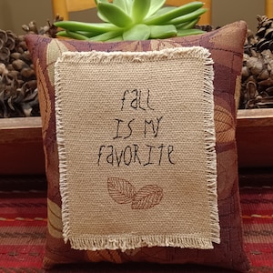 May include: A decorative pillow with a succulent plant on top. The pillow has a burlap patch with the embroidered text "fall IS MY favorite" and two leaf designs. The pillow has a burgundy and gold leaf pattern.