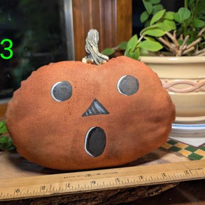 May include: A plush orange pumpkin with a painted black jack-o'-lantern face featuring two circular eyes, a triangular nose, and an oval mouth. A wooden ruler placed in front shows the pumpkin is approximately 20 cm wide.