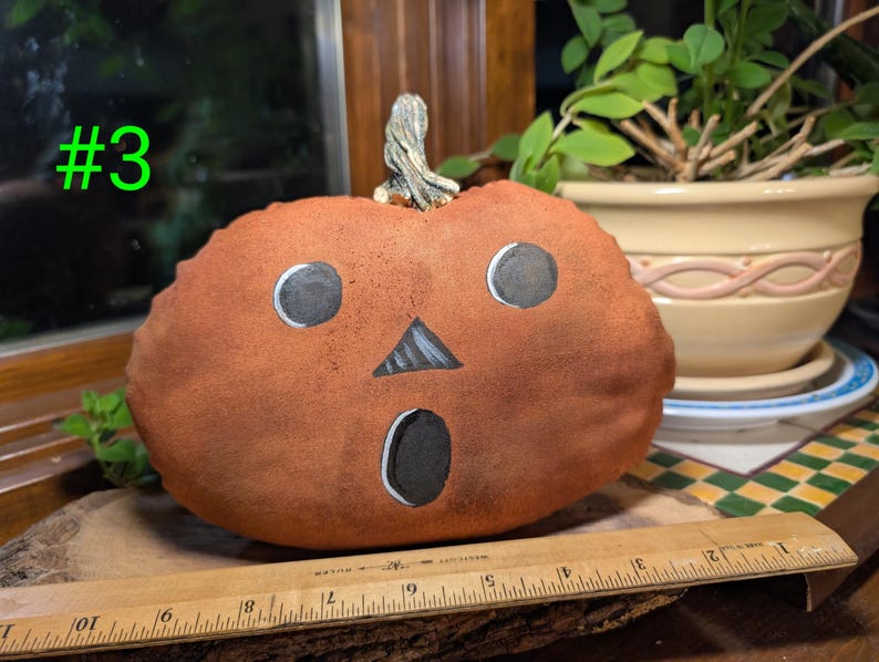May include: A plush orange pumpkin with a painted black jack-o'-lantern face featuring two circular eyes, a triangular nose, and an oval mouth. A wooden ruler placed in front shows the pumpkin is approximately 20 cm wide.