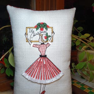 May include: A rectangular off-white textured fabric cushion featuring an embroidered illustration of a person in a red and white striped dress holding a sign that reads It's Christmas. A wooden ruler is visible at the bottom edge of the cushion.
