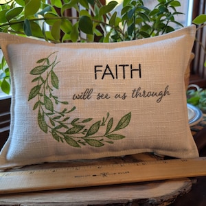 May include: Rectangular beige pillow with the words "FAITH will see us through" in black script. A green leafy design arcs around the left side of the text. The pillow is on a wooden surface.
