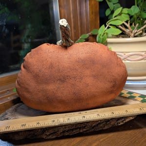 May include: A terracotta-coloured fabric pumpkin with a textured surface and a realistic stem. A wooden ruler is placed in front of the pumpkin, showing it measures approximately 25.4 cm in width.