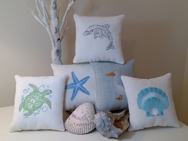 Peut inclure: Plusieurs coussins d&eacute;coratifs carr&eacute;s et rectangulaires avec des motifs brod&eacute;s sur le th&egrave;me de la mer. Les coussins pr&eacute;sentent des images de dauphin, d'&eacute;toile de mer, de tortue, de p&eacute;lican et de coquillage en bleu et vert. Des coquillages sont &eacute;galement inclus.
